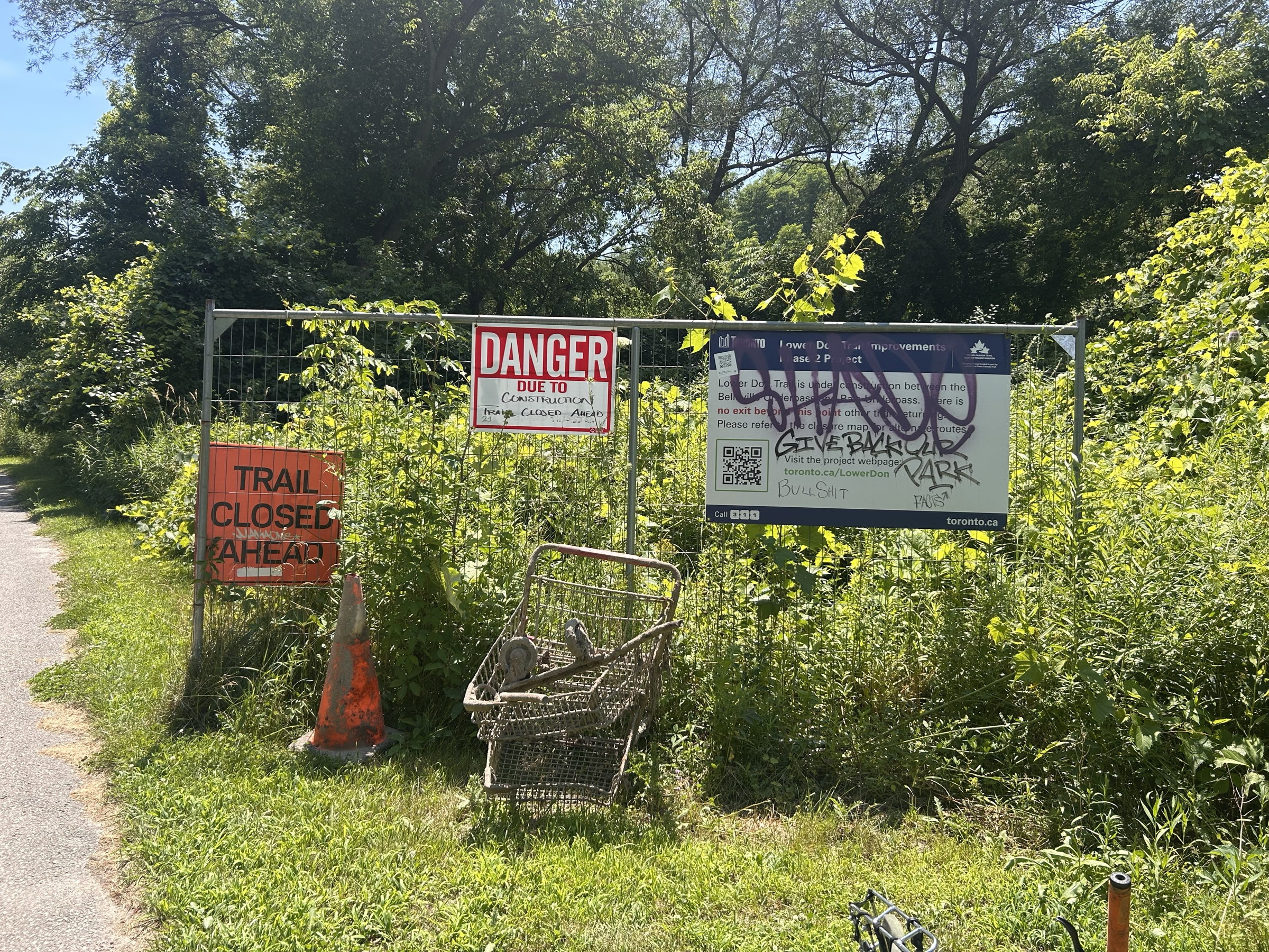 A fenced-off section of the Lower Don Trail: a red Trail Closed Ahead sign, a Danger notice, a construction poster covered in graffiti, an abandoned shopping cart in the weeds