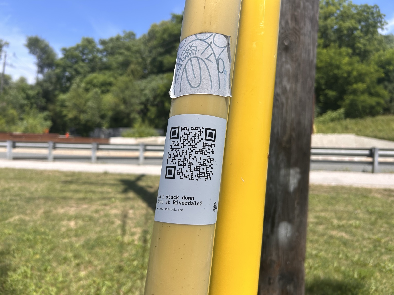 No Road Block sticker asking 'Am I stuck down here at Riverdale?' on a yellow bollard beside a wooden pole