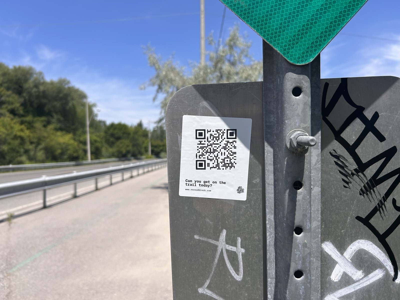 No Road Block sticker asking 'Can you get on the trail today?' on the back of a galvanised road sign beside a guardrail