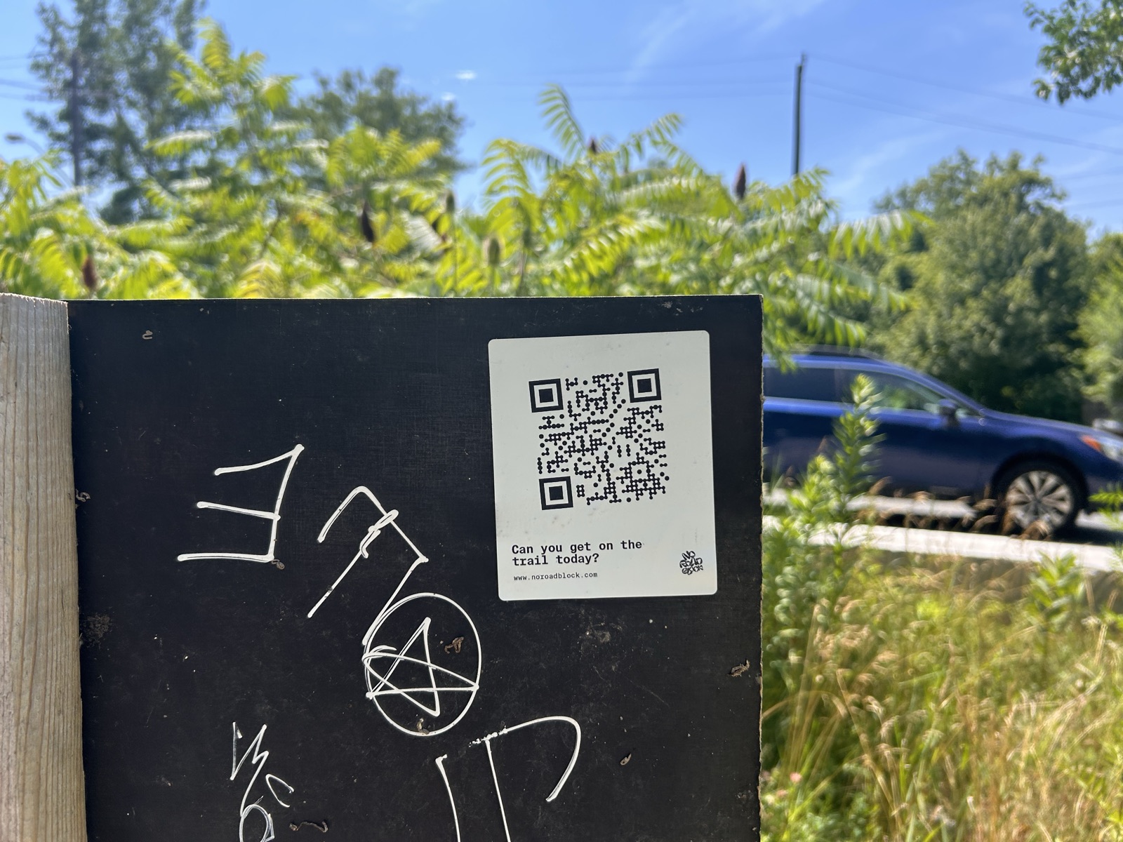 Wider view of the trailhead board with a No Road Block sticker, roadside greenery and a passing car behind